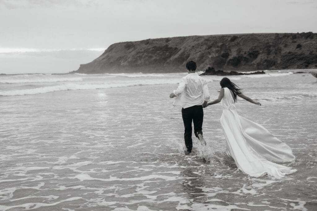 Luxury beach elopement. A black and white image where the bride and groom run into the ocean fully dressed in their wedding attire, her wedding dress catches the waves